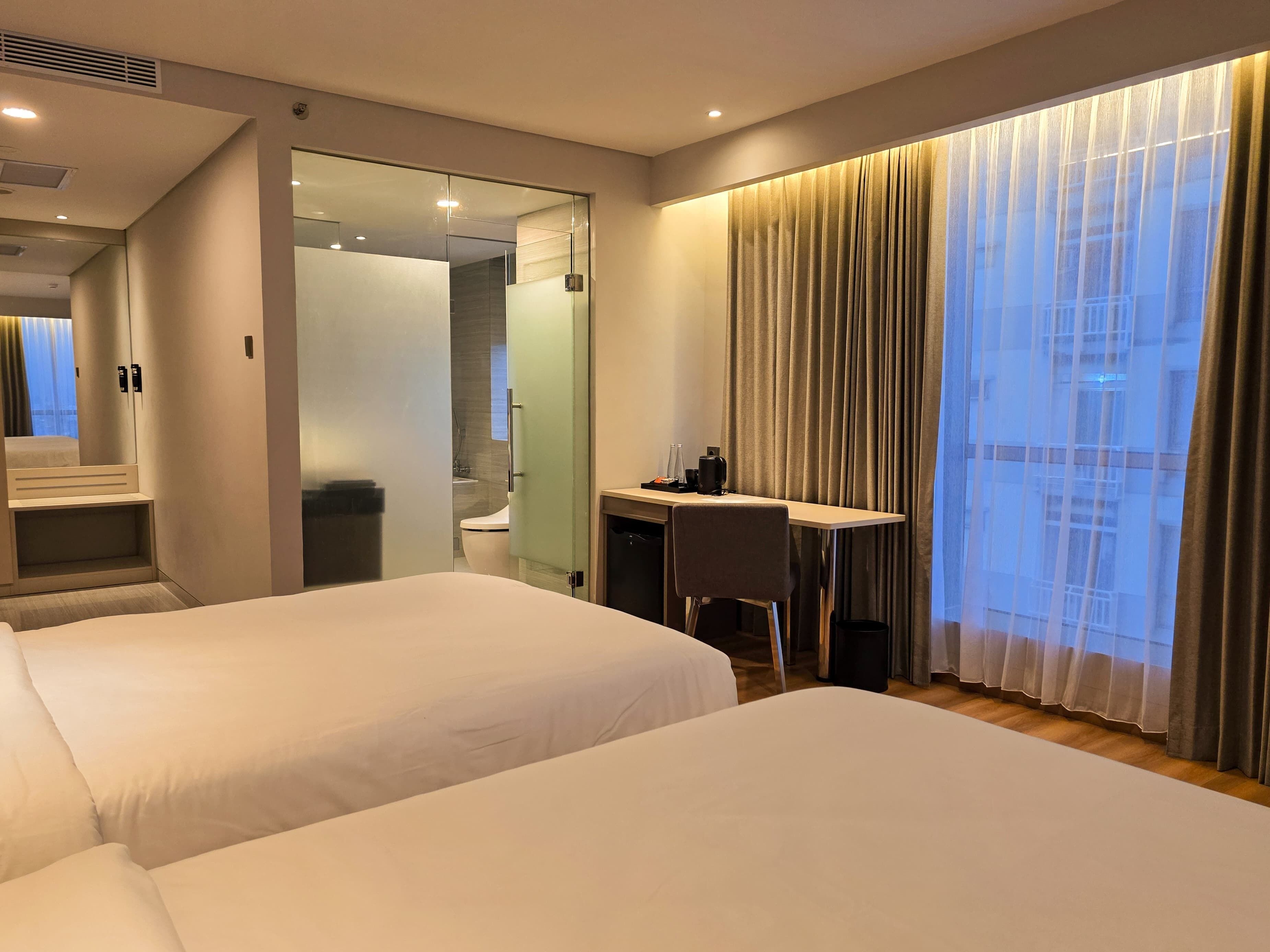 A hotel room with two beds, a desk, a chair, and a large window with curtains.