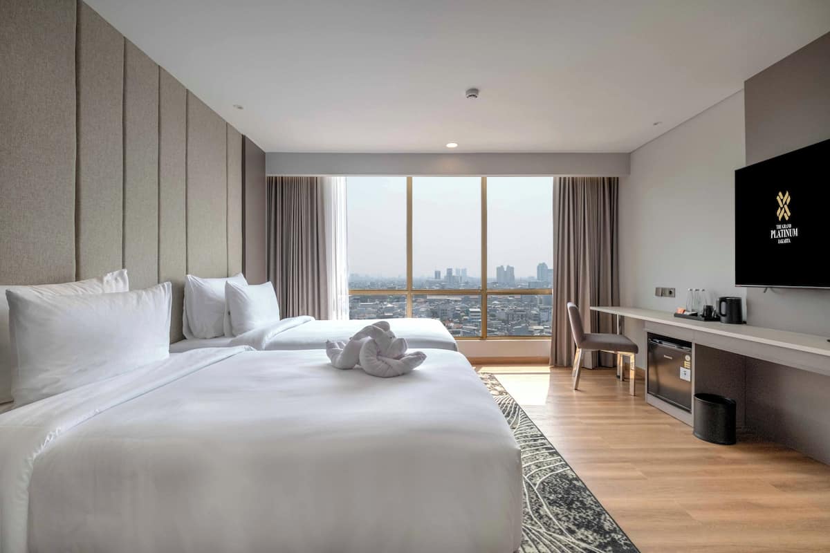 A hotel room with two beds, a desk, a TV, and a city view.
