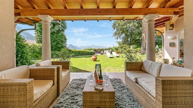 A covered outdoor seating area with wicker furniture and a table with a vase and glasses. - Villa Tre Pini Garden Escape (Lugano)