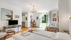 A hotel room with two beds, a desk with a television, a chandelier, and a view of the outdoors. - Villa Tre Pini Garden Escape (Lugano)