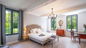 A bedroom with a large bed, a chandelier, a wooden desk, and a view of greenery through the windows. - Gite Les Bons Amis Fronsac 100m2 - 3ch - 6pax - Piscine chauffée - jardin 1300m2 (Fronsac)