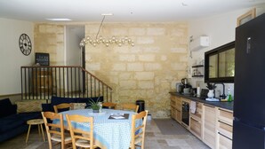 A dining area with a round table, chairs, and a chandelier. A kitchen with a refrigerator and stove is visible in the background. - Gite Les Bons Amis Fronsac 100m2 - 3ch - 6pax - Piscine chauffée - jardin 1300m2 (Fronsac)