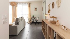 A living room with a sofa, a small table with chairs, a decorative wall hanging, and a shelf with a pineapple and a lamp. - Charming Apartment  2BR/4P - Near to Lyon (Caluire-et-Cuire)