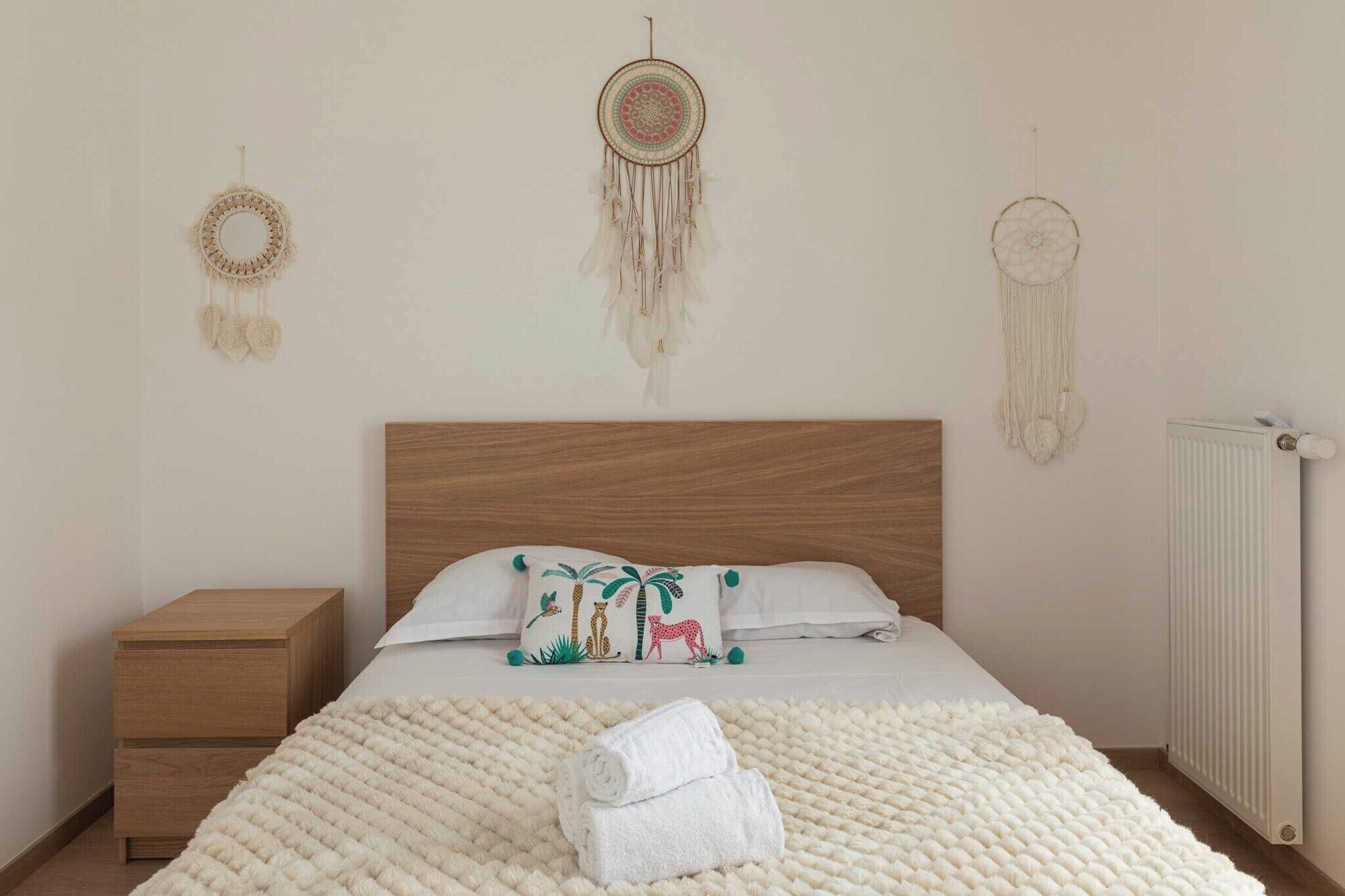 Une chambre avec une tête de lit en bois, une couvre-lit blanche, un coussin décoratif à motifs animaliers et un attrape-rêves accroché au mur.