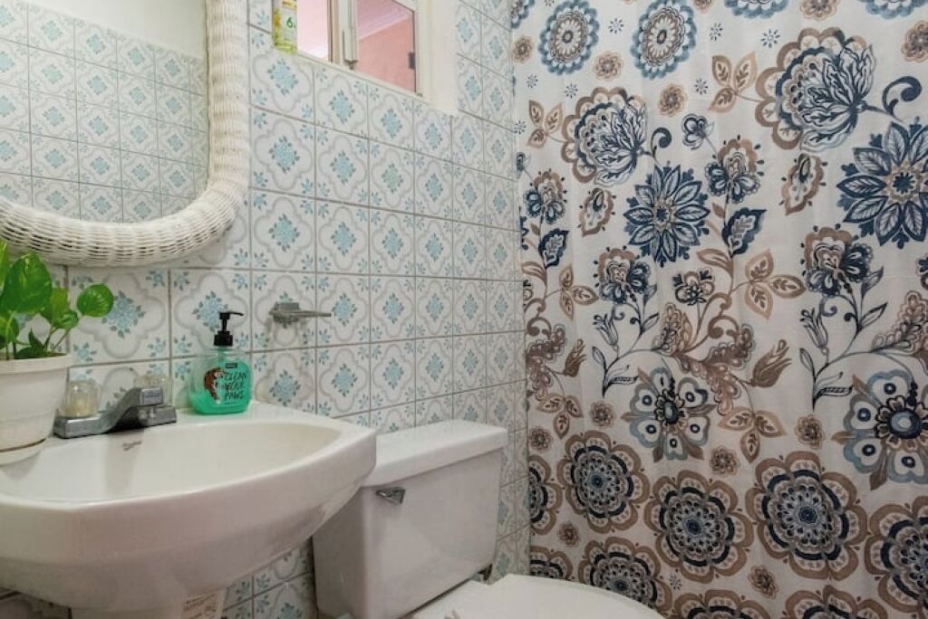 Un baño con un lavabo blanco, un inodoro y un espejo. Hay una planta en maceta y una botella de jabón para manos sobre el lavabo.