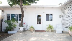 A white building with a gravel driveway, a door, and two windows. There are potted plants and a chair outside.