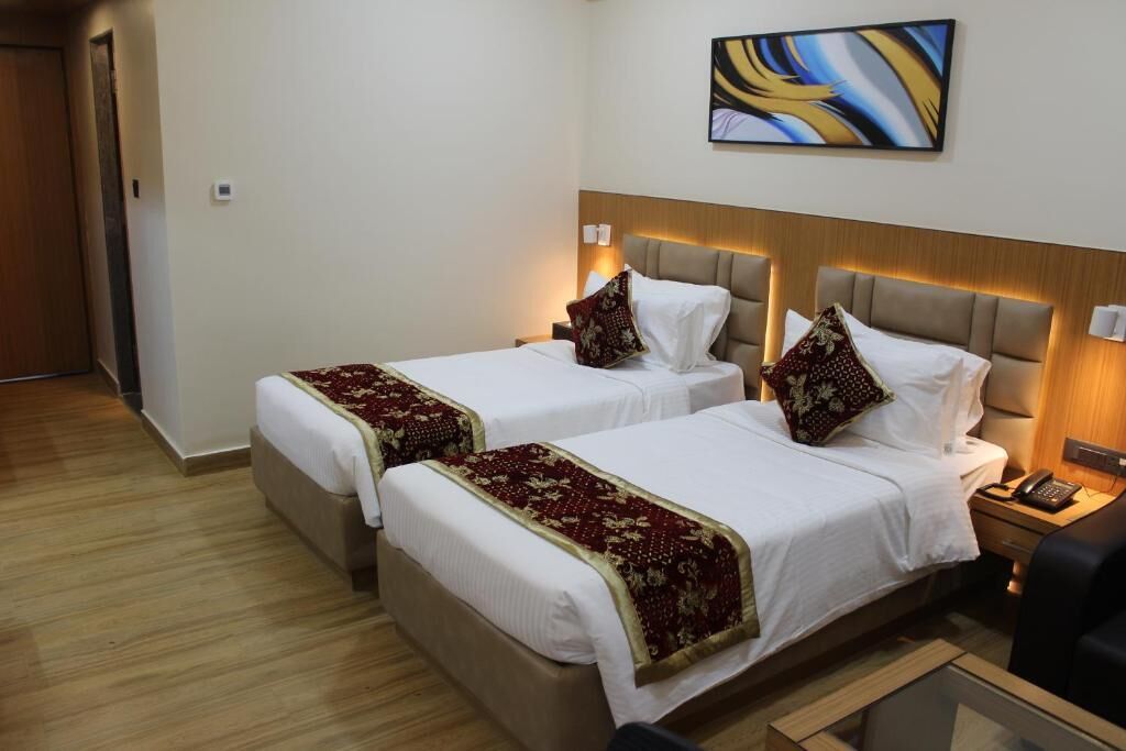 A hotel room with two beds, each with a headboard and bedside tables. There is a painting on the wall above the beds.