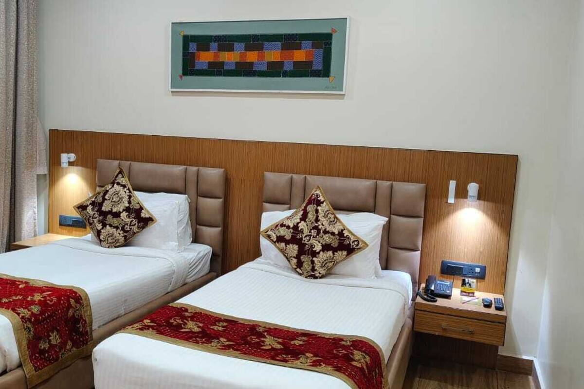 A hotel room with two beds, each with a red and gold bedspread, a wooden headboard, and a nightstand with a phone and remote controls.