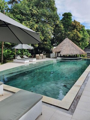 Ein Pool mit Liegestühlen und Sonnenschirmen, umgeben von üppiger Vegetation und einem Gebäude mit einem Reetdach.