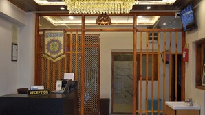 Uma área de recepção com balcão identificado como "RECEPTION", um candelabro, uma televisão fixada na parede e um biombô decorado com caligrafia árabe.