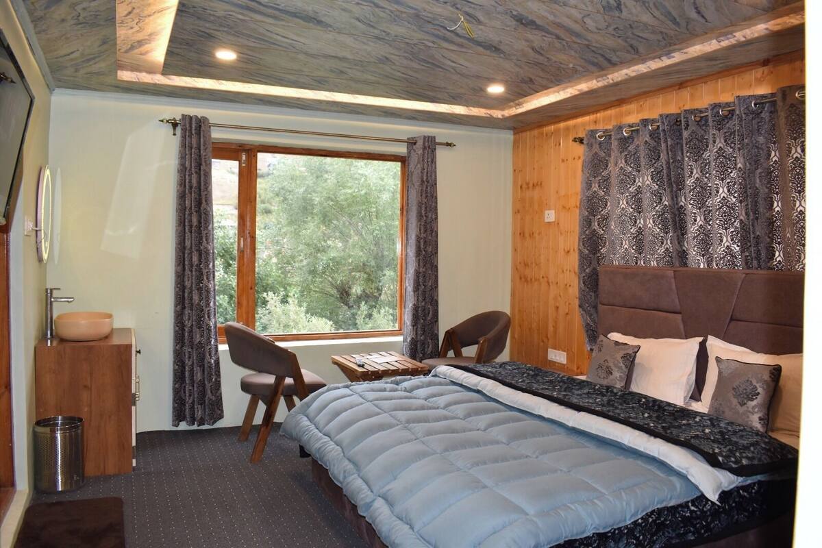 A bedroom with a bed, a desk, and a chair. There is a window with curtains, a wooden headboard, and a small cabinet with a bowl.