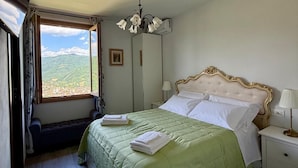 A bedroom with a bed, a nightstand, a lamp, a window with a view of mountains, and a ceiling fan. - Charming Guest Room in Villa Casavecchia with Mountain Views (Dicomano)