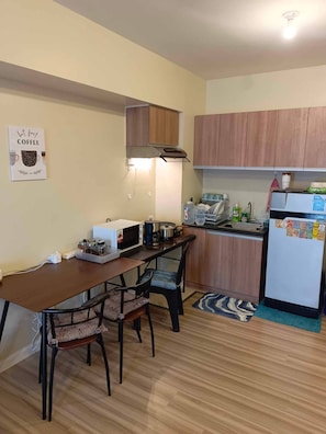 A compact kitchen with wooden cabinets, a dining table with chairs, a microwave, a coffee maker, and a refrigerator.