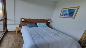A bedroom with a wooden headboard, a bed with white bedding, a small bedside table with a lamp, and a framed picture on the wall. - Modern Mountain Apartment with Balcony (Chamrousse)