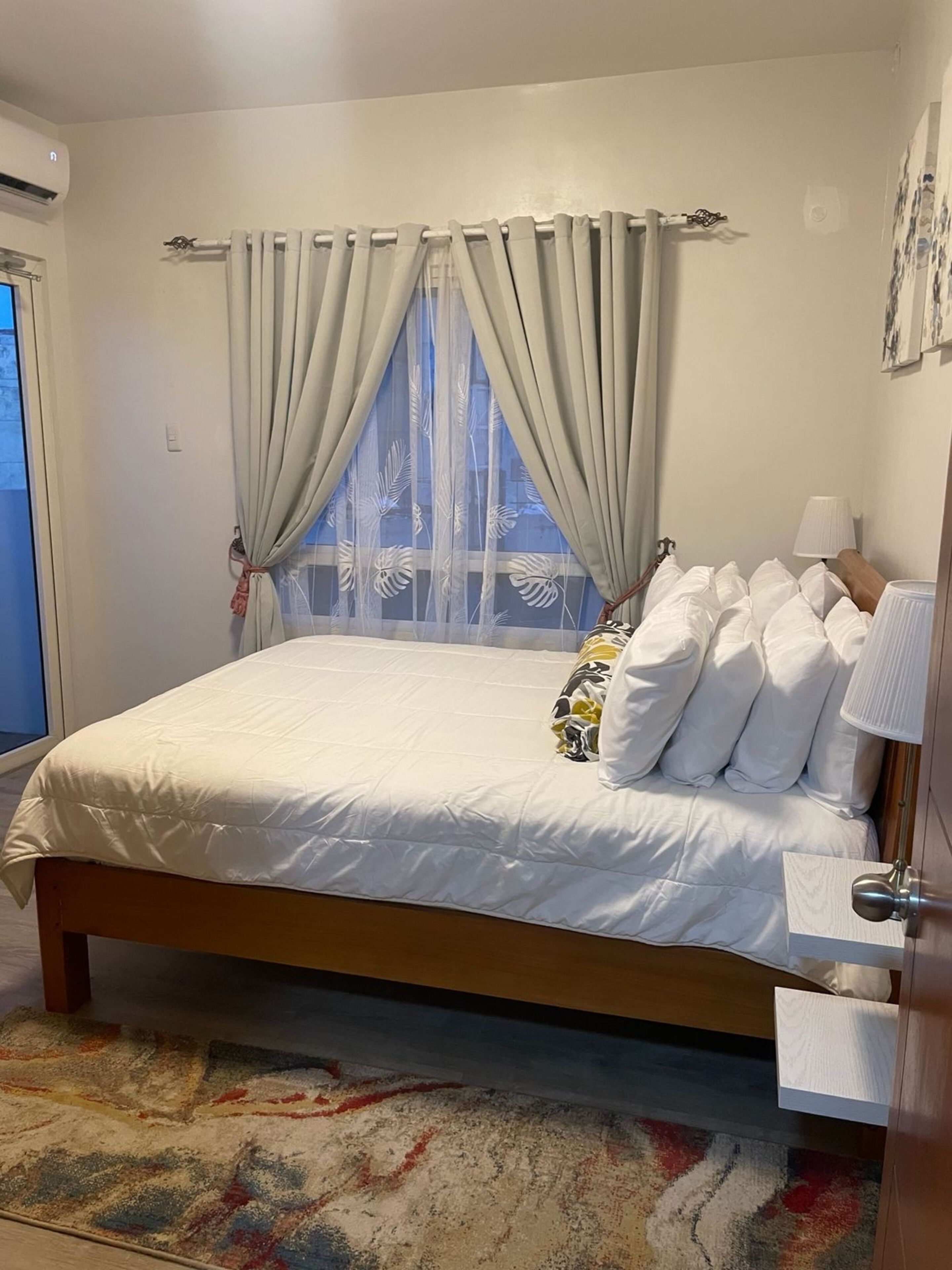 A neatly made bed with white bedding and a floral pillow, a bedside lamp, and a window with sheer curtains.