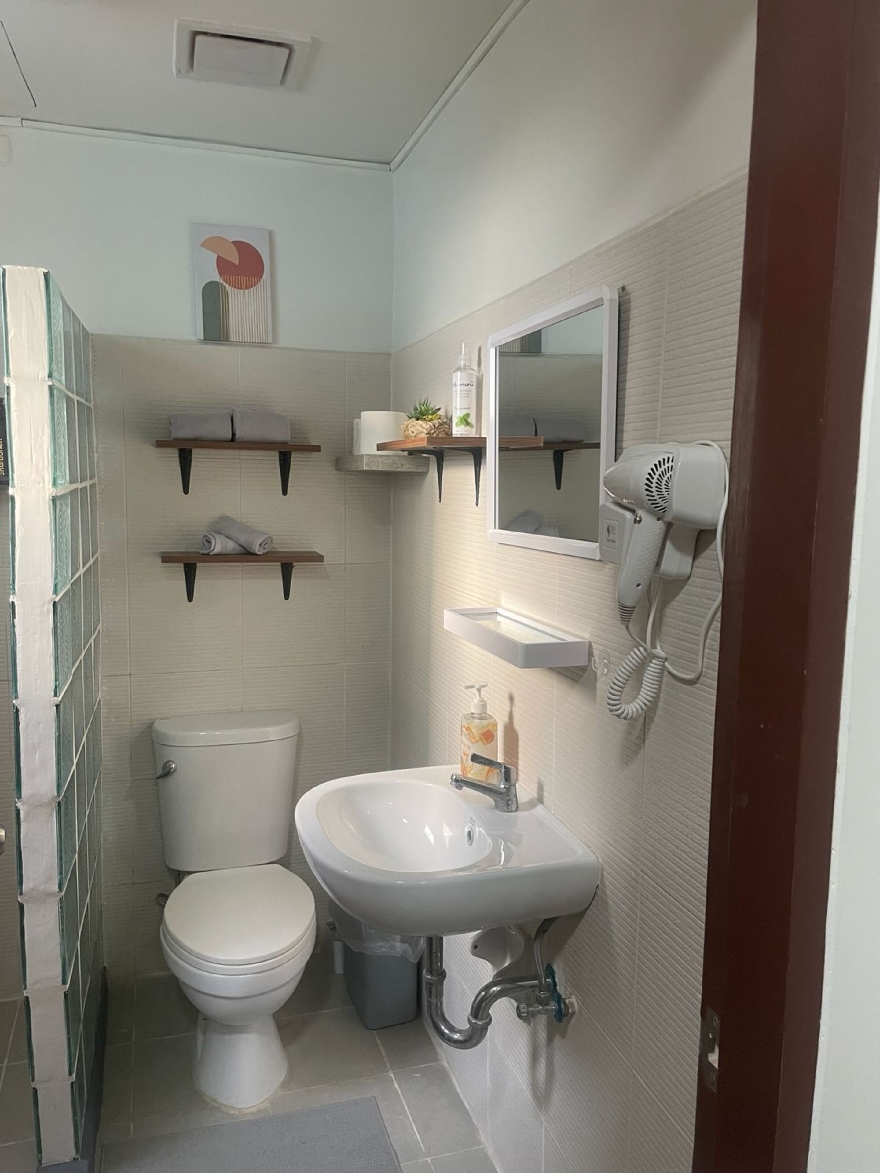 Un baño con inodoro, lavabo y secador de pelo en la pared. Hay repisas sobre el lavabo.