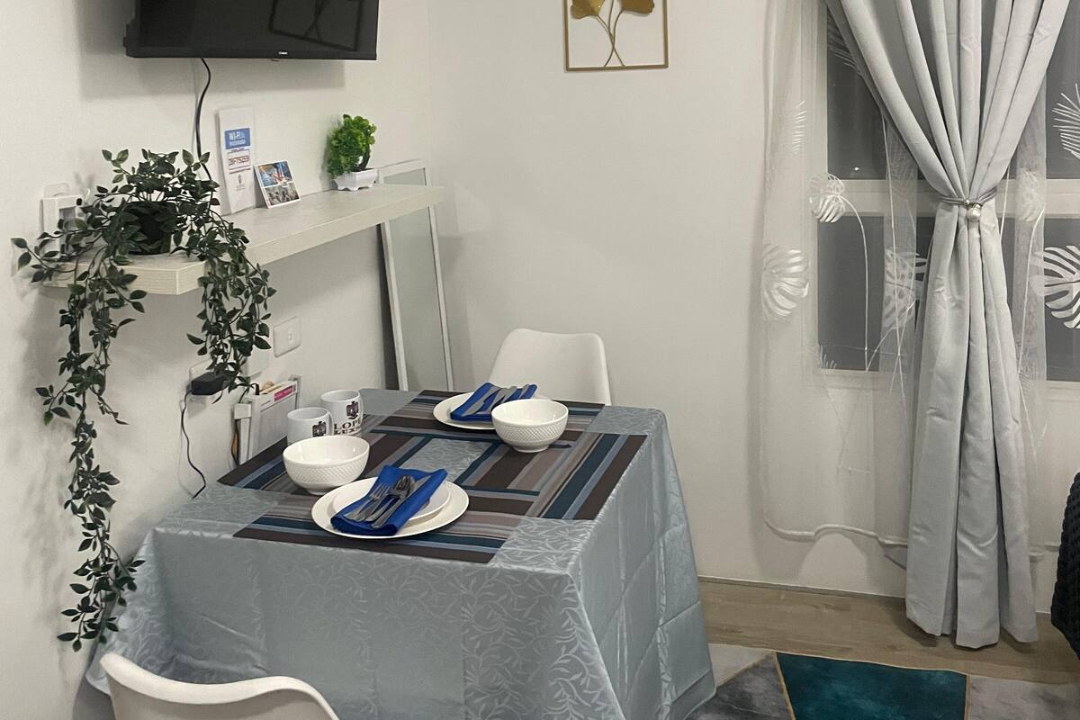 A dining area with a table set for two, a television mounted on the wall, and a window with curtains.