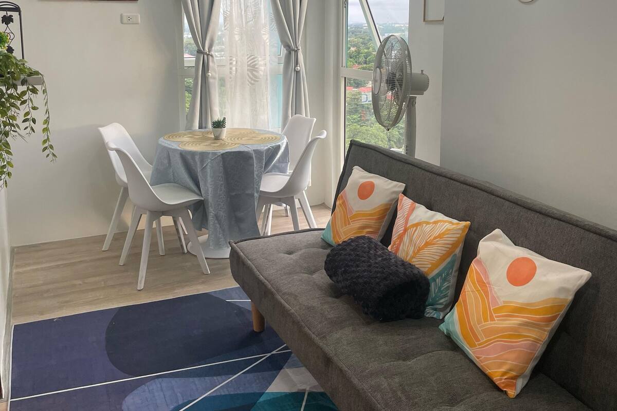 A modern living room with a grey sofa, colorful cushions, a dining table with chairs, a large window with curtains, and a ceiling light fixture.