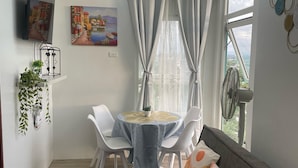 Un salon moderne avec un canapé gris, une table à manger avec des chaises, un téléviseur fixé au mur et une grande fenêtre avec des rideaux.