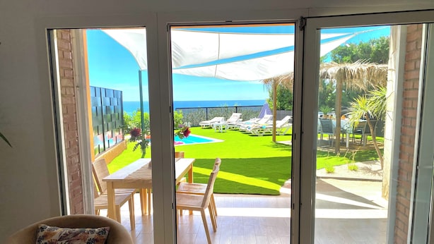 Vista desde una habitación con puerta corrediza de vidrio a un espacio exterior con piscina, sillones de descanso y césped.
