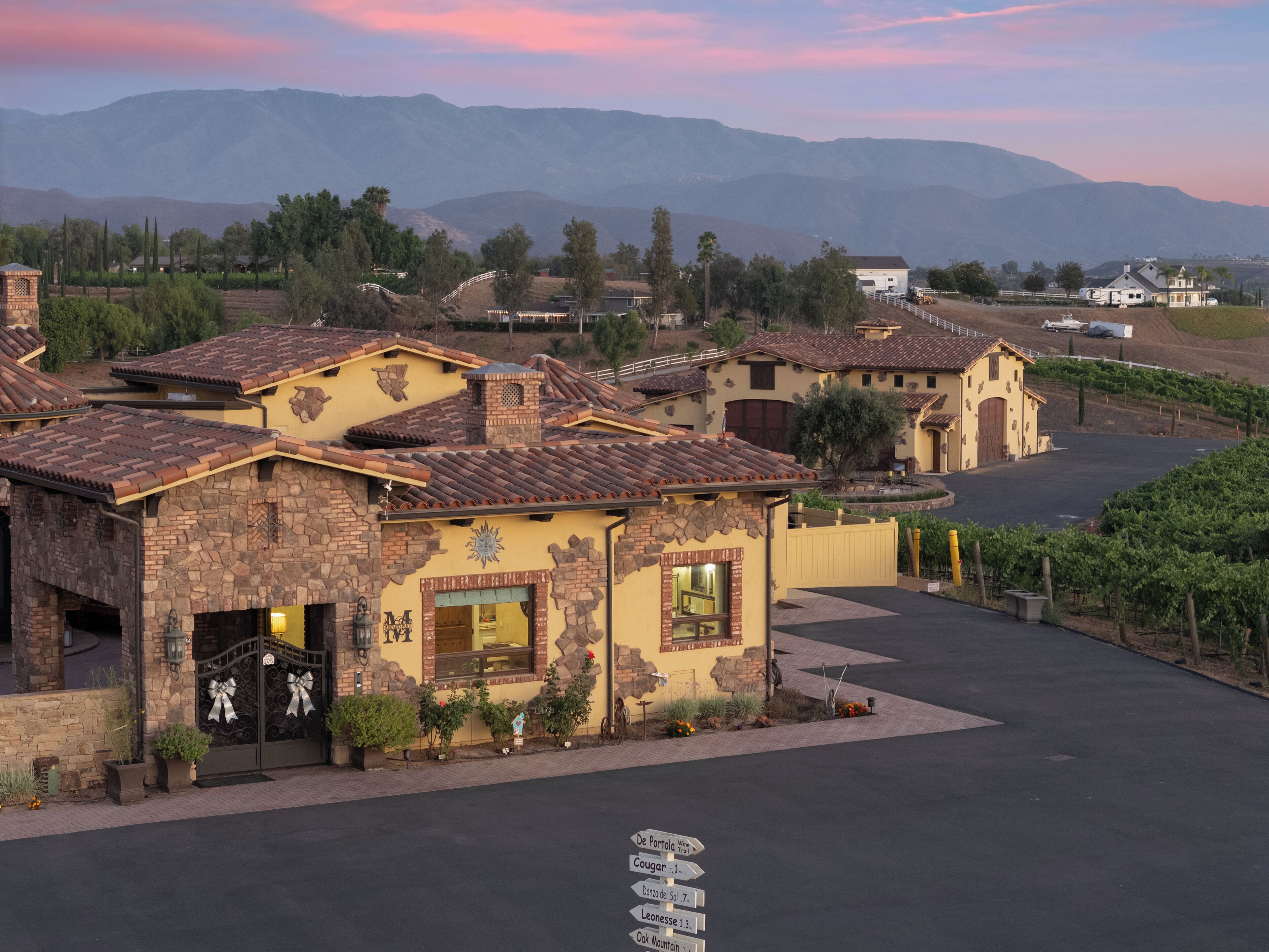 A residential area with houses featuring stone and stucco exteriors, a paved driveway, and a vineyard in the background.