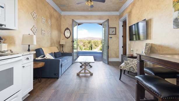 A spacious living area with a sofa, coffee table, and a dining table with chairs. A ceiling fan is visible, and there is a view through an open door to the outdoors. - New-Build Temecula Villa with King Suite, Patio & Vineyard Views | The Villa (Temecula)