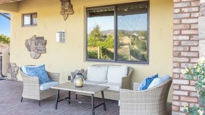 An outdoor patio with wicker furniture, a table, and a view of a vineyard through a large window. - New-Build Temecula Villa with King Suite, Patio & Vineyard Views | The Villa (Temecula)