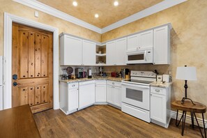 Une cuisine avec des meubles blancs, une porte en bois, un four à micro-ondes, un four traditionnel et une petite table avec une lampe.