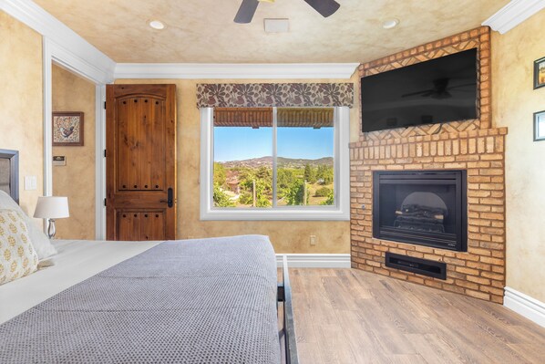 A bedroom with a large window, a fireplace, and a television mounted on the wall. - New-Build Temecula Villa with King Suite, Patio & Vineyard Views | The Villa (Temecula)