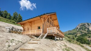 A wooden cabin with a balcony, situated on a hillside with a mountain in the background.