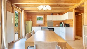 A wooden cabin with a dining table, chairs, a kitchen with white cabinets, and a window overlooking greenery.