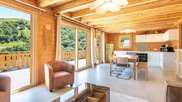 A spacious wooden cabin with large windows offering a view of a lush green landscape. Inside, there is a cozy seating area with a leather sofa and a dining table.