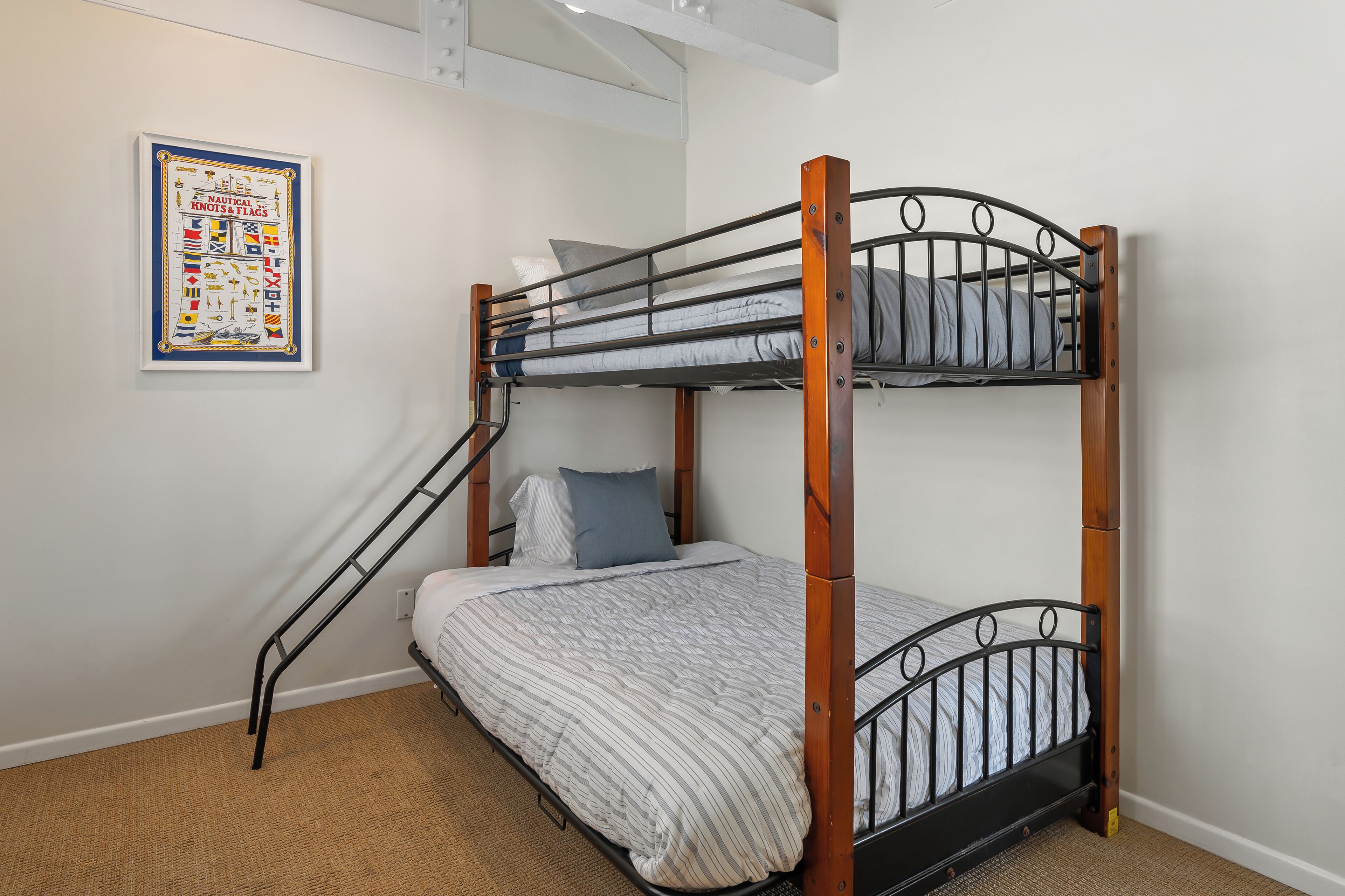 Una cama litera con estructura de madera y barandillas metálicas, una cama individual con una colcha a rayas, y un póster con temática náutica en la pared.