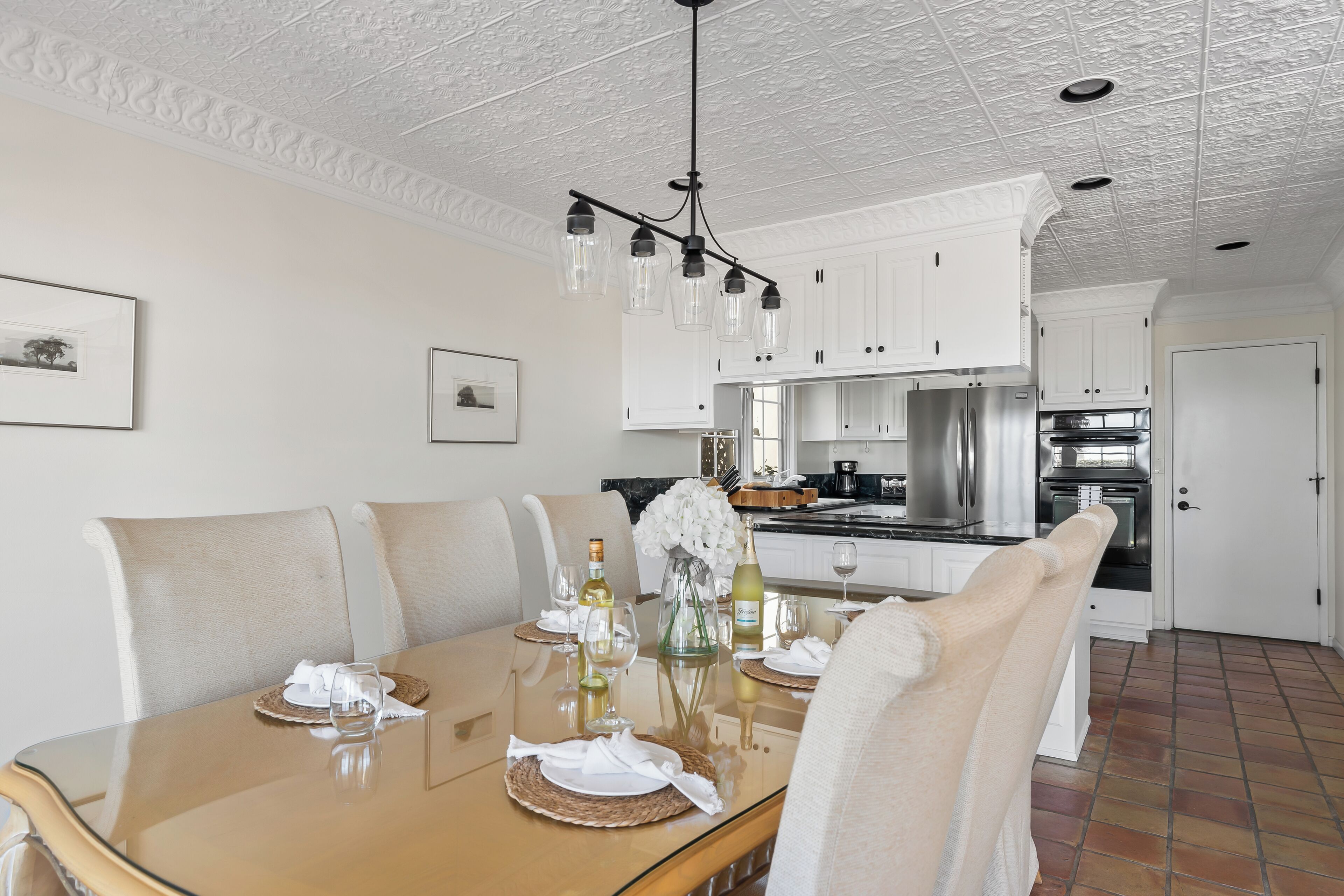 A dining area with a glass table, white chairs, and a chandelier. There are framed pictures on the wall and a kitchen in the background.