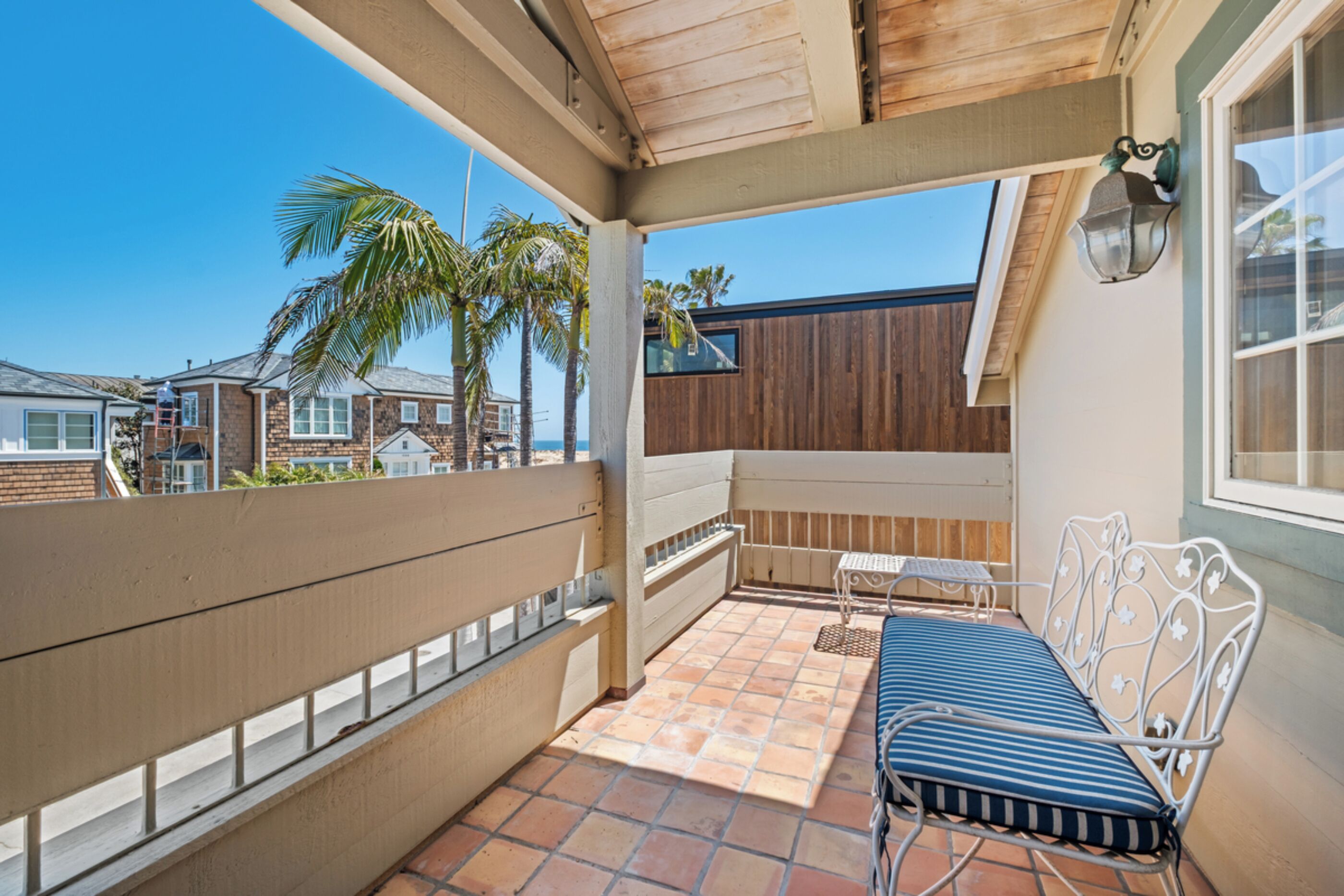 A covered balcony with a metal bench, a small table, and a view of palm trees and neighboring houses.