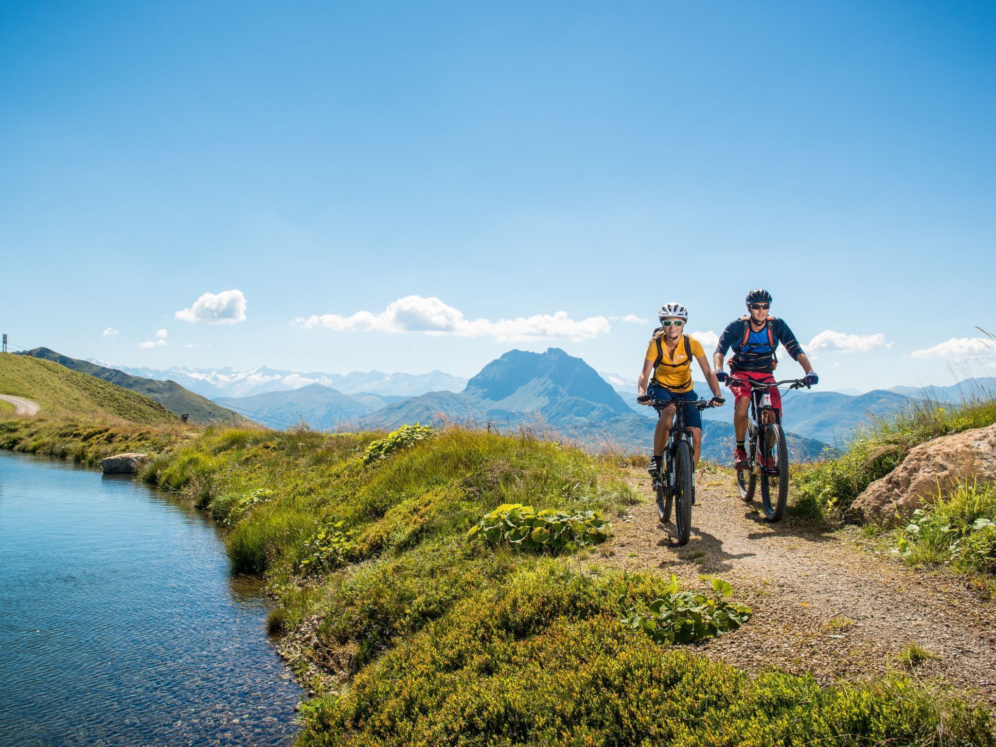 Two cyclists on a mountain path with a lake and mountains in the background.