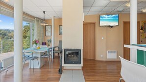 A spacious room with a fireplace, a dining area with a table and chairs, a television mounted on the wall, and a view of the outdoors through large windows.