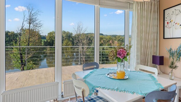 A dining area with a table set for two, a vase with flowers, and a view of a lake and trees through large windows.