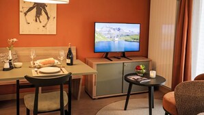 A cozy hotel room with a dining area, a television displaying a scenic landscape, and a small table with a candle and a magazine. - Juliana by Interhome (Zermatt)