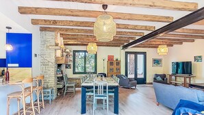 Zona d'estar àmplia amb taula de menjador, cadires i sofà. Hi ha una escala que mena a un altre pis, i una prestatgeria plena de llibres.