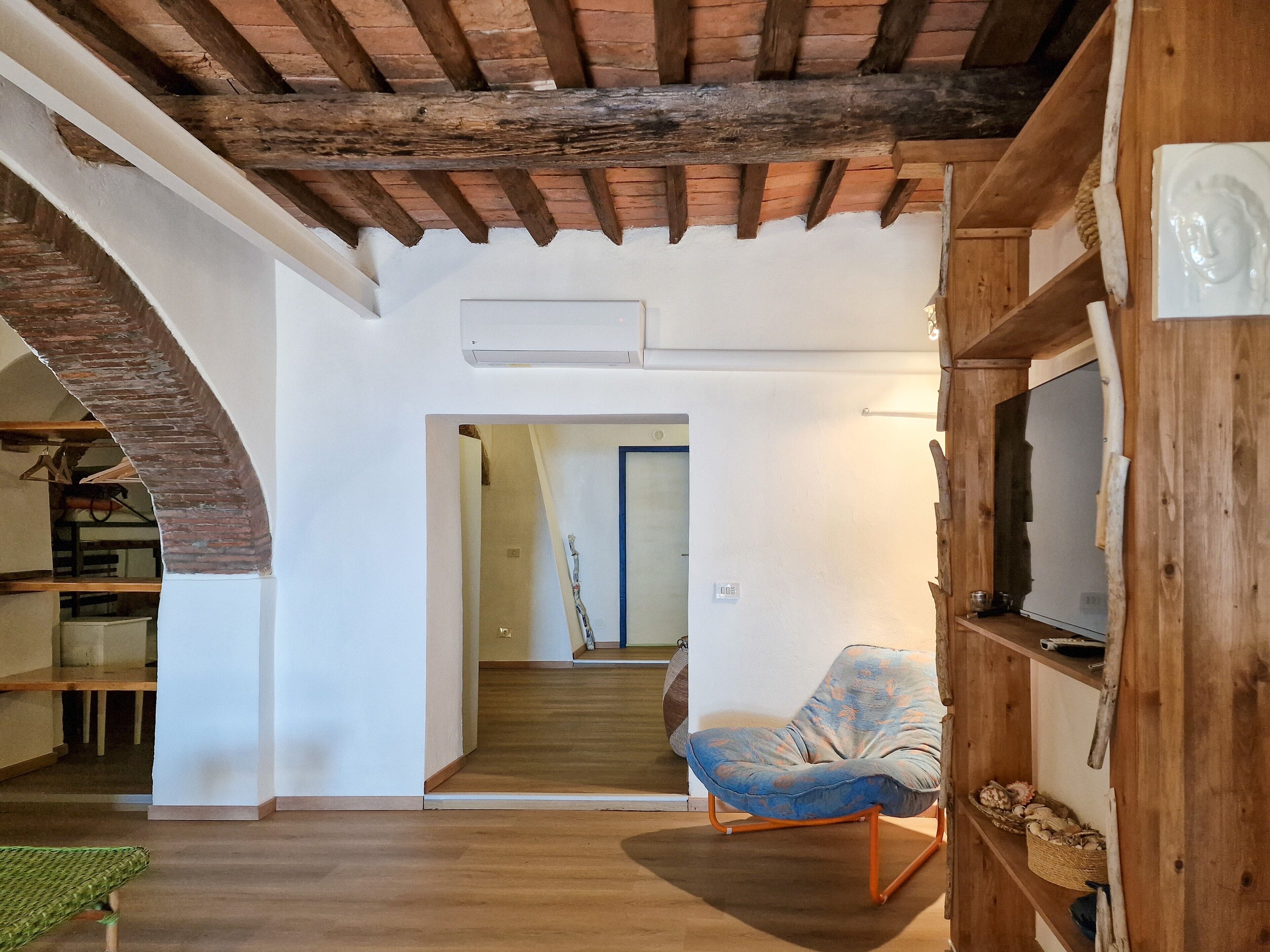 Een kamer met een houten plafond, een televisie op een houten standaard, een blauwe stoel en een trap naar een andere kamer.