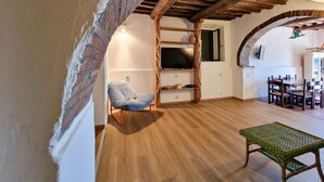 Un interior rústico con arcos de ladrillo visto, vigas de madera, un televisor, una mesa de comedor con sillas y un banco de mimbre.