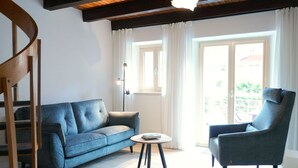 A living room with a blue sofa, a round table, a chair, a wooden staircase, and a large window with curtains. - Rustico Emilio by Interhome (Roveredo)