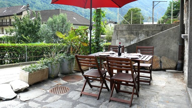 An outdoor patio with a table and chairs, a red umbrella, and a view of mountains in the background. - Rustico Emilio by Interhome (Roveredo)
