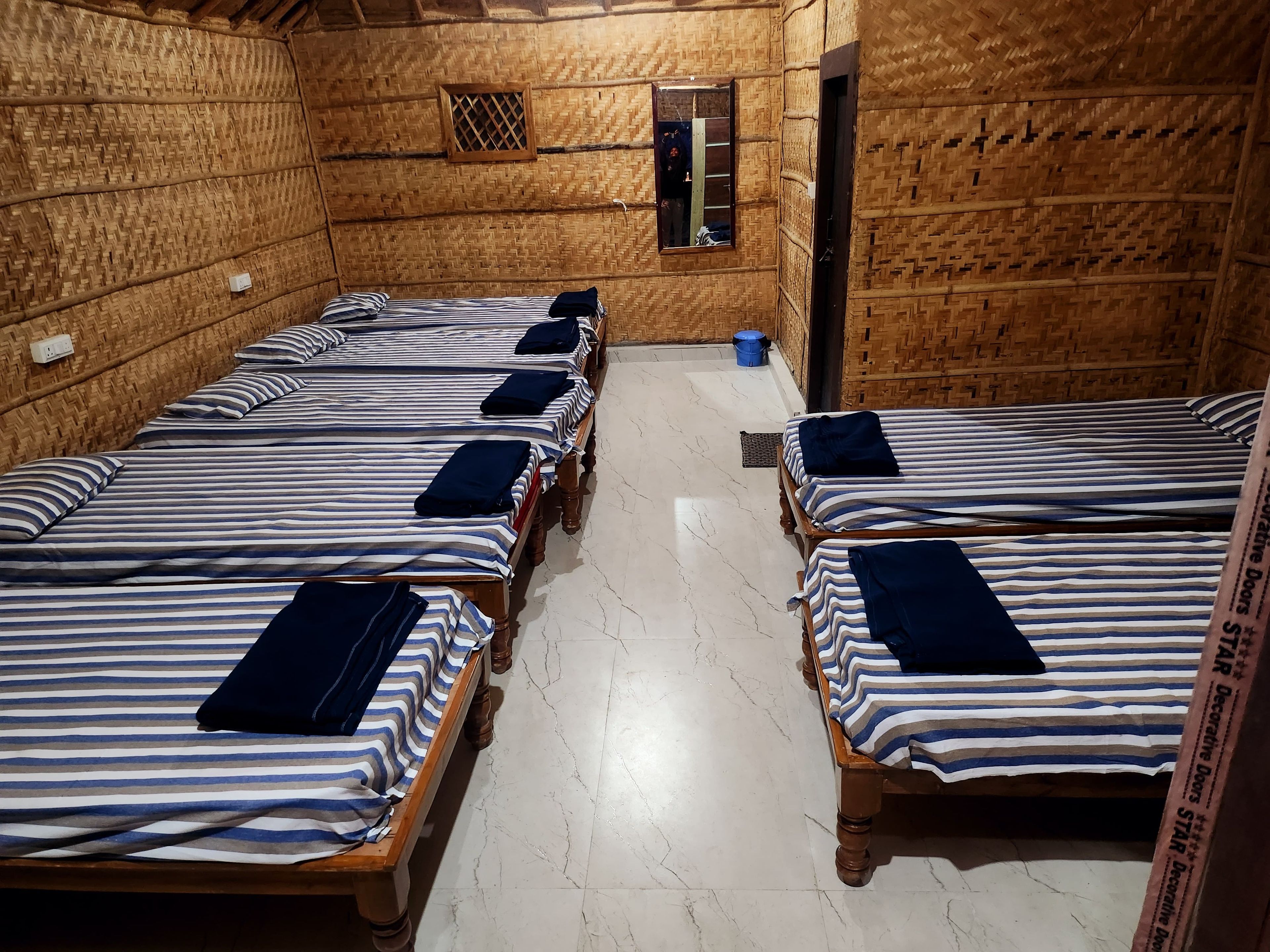 Una habitación con cuatro camas de madera, cada una con ropa de cama a rayas y almohadas oscuras. El piso es de baldosas y hay una ventana pequeña con un diseño de cuadrícula.