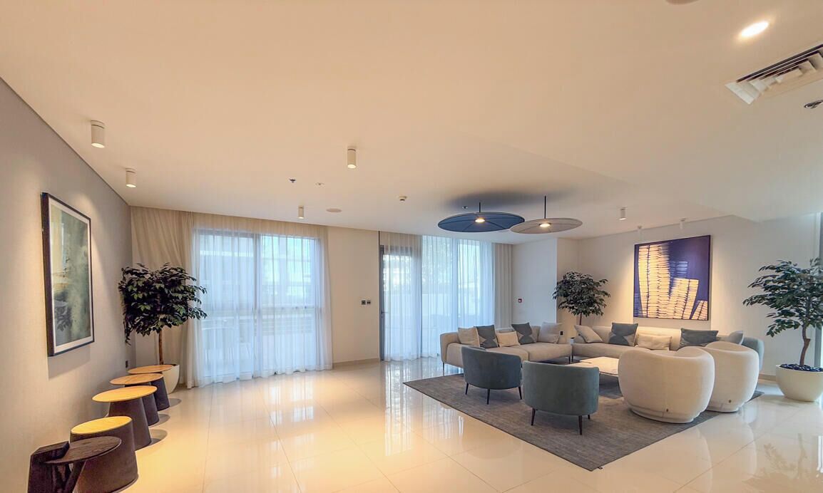 A spacious living room with a modern sofa set, a coffee table, and a large window with sheer curtains. There are potted plants and a framed artwork on the walls.