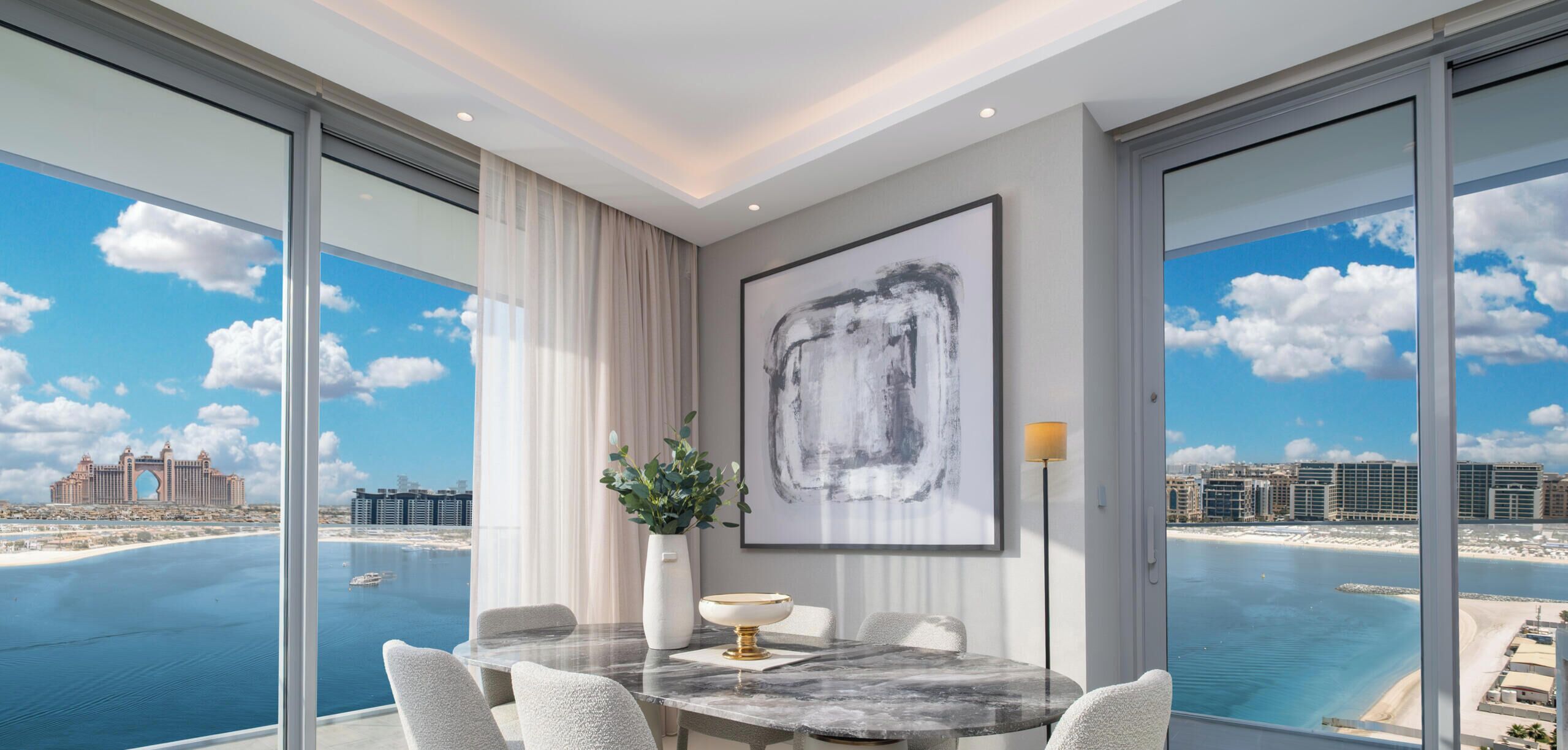 A modern dining area with a marble table, white chairs, and a large window offering a view of the sea and cityscape.