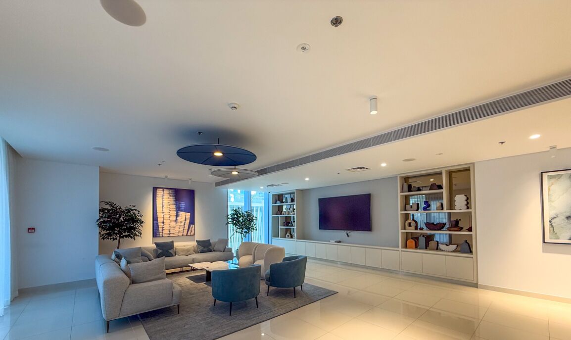 A modern living room with a sofa set, a coffee table, and two armchairs. There is a bookshelf with decorative items and a flat-screen TV mounted on the wall.