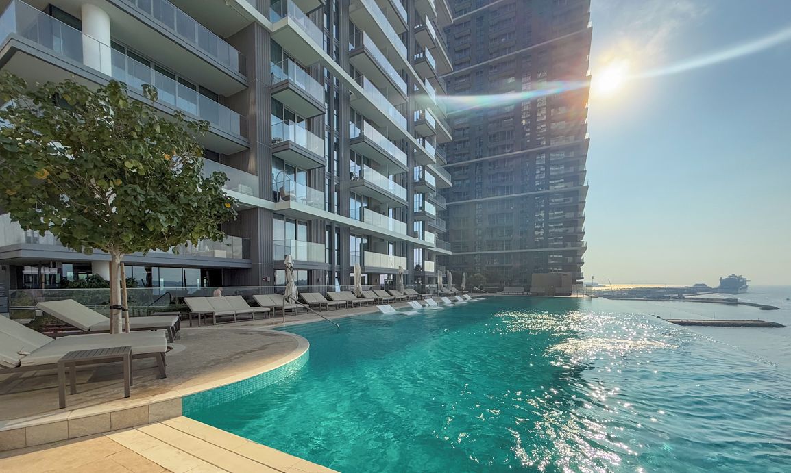 A modern hotel with a poolside area, a large building in the background, and a clear view of the sea.