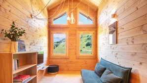A cozy cabin with wooden walls, a loft bed, a sofa, a bookshelf, and two windows offering a view of a green landscape.